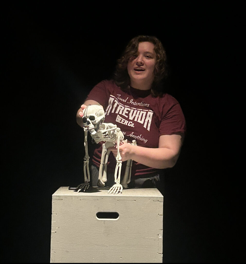 Sam has shoulder length brown wavy hair. They are wearing a maroon shirt and holding a skeleton, positioned to look like a cow.