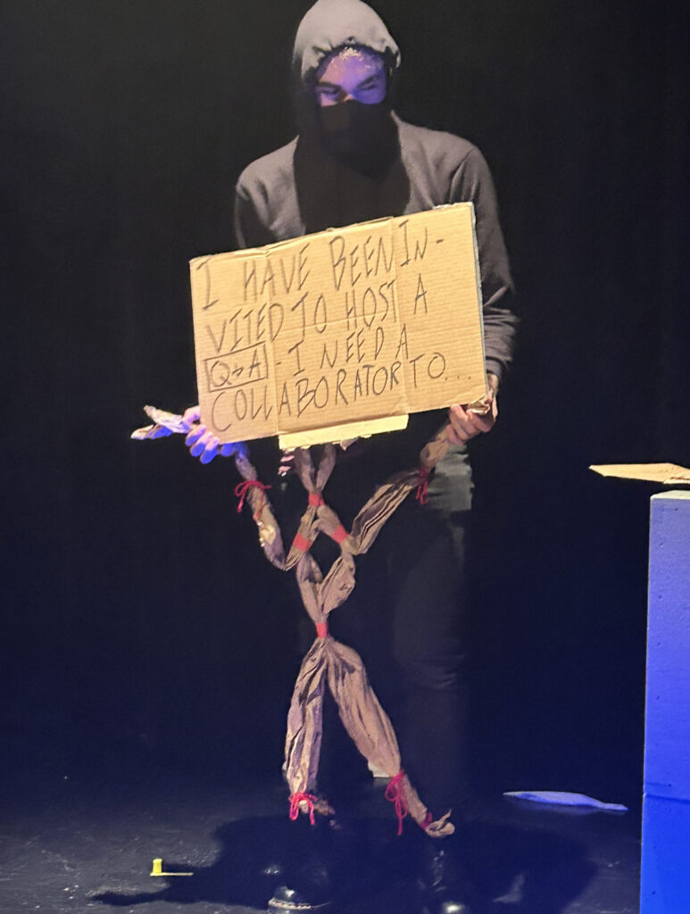 Nick is wearing a black hoodie and has face mask on. He is holding a cardboard sign that reads "I have been invited to host a Q&A - I need a collaborator to..."